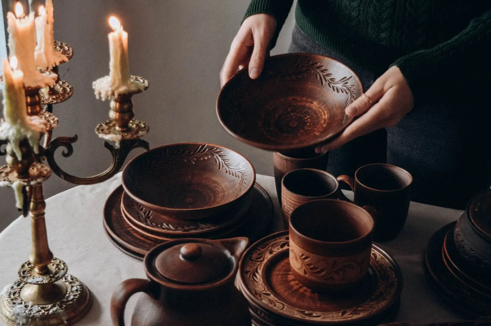 Hands setting a candlelit table with handmade clay bowls, cups and plates